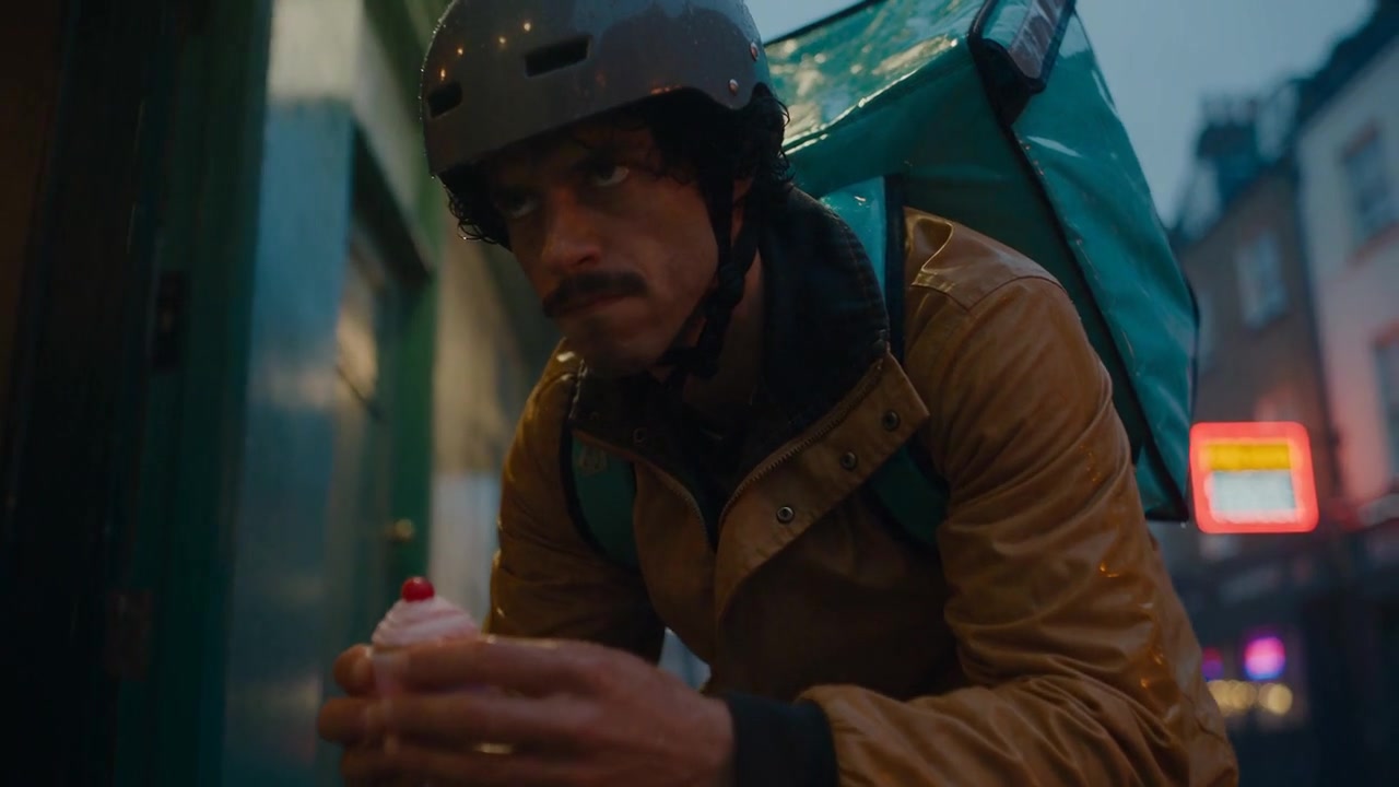 A courier holds a finished cupcake on a wet street at dusk.